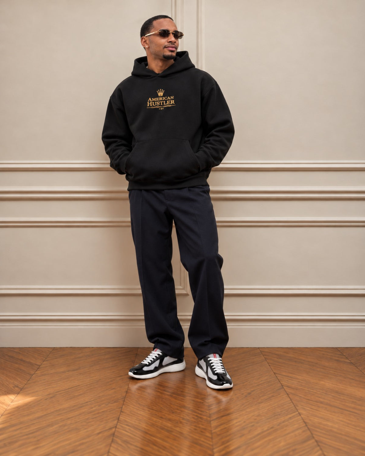 Ryan wearing the black American Hustler signature 500 GSM hoodie, relaxed dark trousers and black and silver sneakers against a warm paneled wall.
