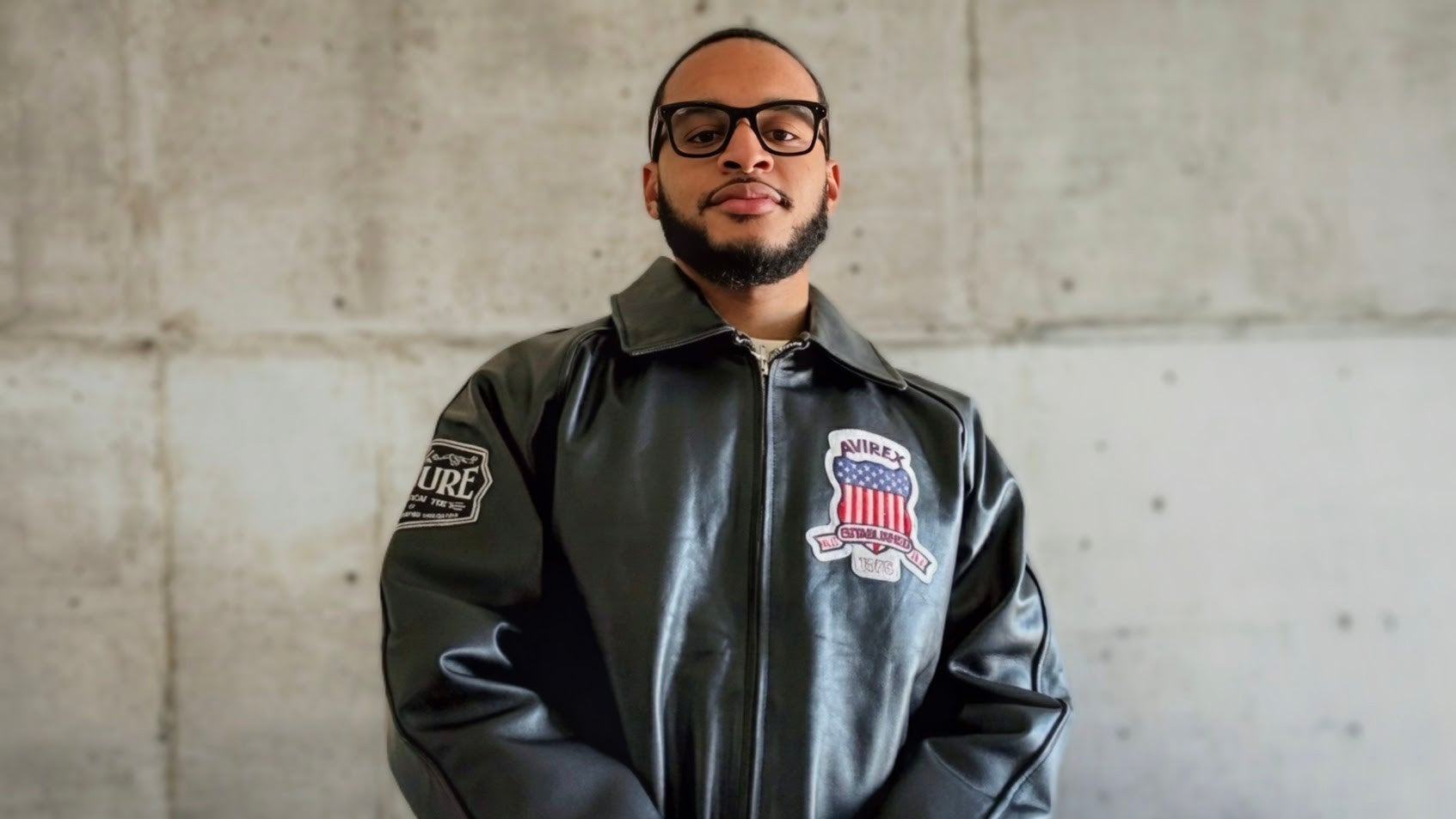 Man wearing a black Culture Limited Tee Icon leather jacket with patches against a concrete walle